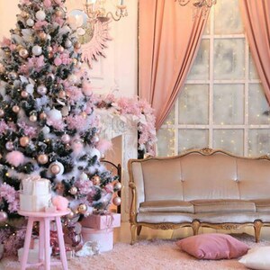 Pode incluir: Uma árvore de Natal rosa e branca decorada com enfeites, luzes e neve artificial. Um sofá rosa com detalhes dourados fica em frente a uma janela com cortinas transparentes.