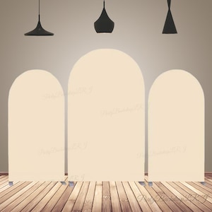 Puede incluir: Tres soportes de telón de fondo de color beige en forma de arco con la parte superior redondeada. Los telones de fondo están hechos de tela y son independientes. Los telones de fondo están instalados en un suelo de madera con tres lámparas colgantes negras que cuelgan del techo.