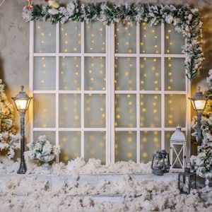 Pode incluir: Um fundo de janela branco com uma guirlanda de vegetação e luzes piscando. Duas árvores de Natal cobertas de neve ladeiam a janela, e há lanternas e outros elementos decorativos no chão nevado.