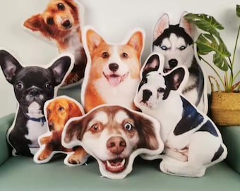 Può includere: Una collezione di cuscini a forma di cane di varie razze, tra cui un bulldog francese, un corgi e un husky. I cuscini presentano immagini realistiche a colori dei volti dei cani, con texture morbide e soffici. I cuscini sono disposti su un divano verde.