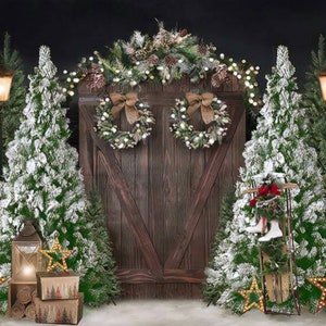 Pode incluir: Uma porta de madeira rústica com uma guirlanda de ramos de pinho e luzes brancas, ladeada por duas árvores de Natal cobertas de neve. A porta é decorada com duas coroas e um laço. A cena é iluminada por luzes de corda e lanternas.