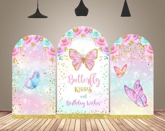 Puede incluir: Tres telones de fondo en forma de arco para una fiesta de cumpleaños. El telón de fondo central es blanco con purpurina dorada y flores rosas. Presenta una mariposa rosa con el texto "I Butterfly Kisses and Birthday Wishes". Los telones de fondo laterales son rosas y azules con purpurina dorada y flores. Cada telón de fondo tiene una mariposa.