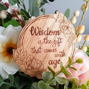 May include: A round wooden sign with a floral design and the text "Wisdom is the gift that comes with age".