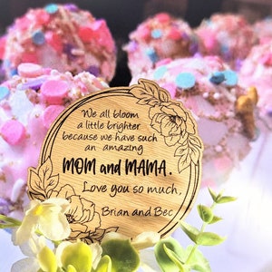 Könnte beinhalten: Eine Holzplakette mit floralen Akzenten und den Worten "Mom and MAMA". Die Plakette ist von weißen und grünen Blumen umgeben und befindet sich vor einem verschwommenen Hintergrund aus rosa und blauen Leckereien. Der Text lautet: "Wir alle erblühen ein wenig heller, weil wir so eine erstaunliche Mom und MAMA haben. Ich liebe euch so sehr, Brian und Bec."