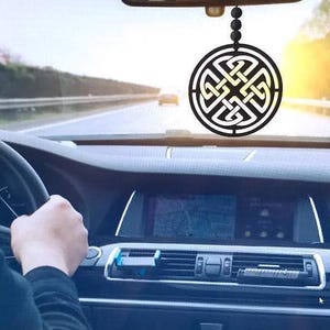 Puede incluir: Un interior de coche negro con un adorno colgante en el espejo retrovisor. El adorno es de diseño circular con un patrón de nudo celta en oro.