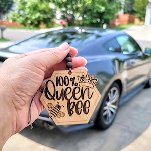 Könnte beinhalten: Ein hölzerner Auto-Lufterfrischer mit dem Text "100% Queen Bee" in Sechseckform, mit einer Krone und einer Bienenillustration. Der Lufterfrischer wird vor einem dunkelgrauen Auto gehalten.