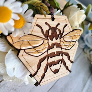 Può includere: Un ornamento di legno a forma di ape esagonale con un designo di ape marrone. L'ornamento è appeso a uno spago bianco ed è circondato da fiori bianchi.