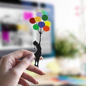 Könnte beinhalten: Ein schwarzer Papierschnitt einer Person, die einen Strauß bunter Ballons hält. Die Ballons sind rot, orange, gelb, grün, blau und lila.