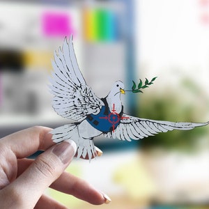 Op de afbeelding: Een witte papieren duif met een blauw en rood doelwit op zijn borst, die een groene olijftak in zijn bek houdt. De duif wordt vastgehouden door een hand.