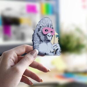 Könnte beinhalten: Eine Hand hält einen Papier-Ausschnitt eines Gorillas, der eine rosafarbene Maske trägt. Der Gorilla ist in Schwarzweiß auf gelbem Hintergrund gezeichnet.