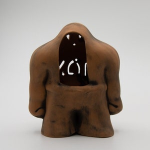 May include: A brown ceramic sculpture of a figure with a large, open mouth and a cutout in the chest area. The figure has a rounded, abstract shape and a textured surface.