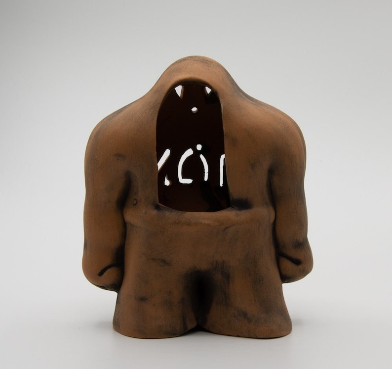 May include: A brown ceramic sculpture of a figure with a large, open mouth and a cutout in the chest area. The figure has a rounded, abstract shape and a textured surface.
