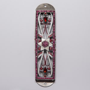 Op de afbeelding: Een zilveren Torah-wijzer met een roze en wit emaille ontwerp. De wijzer heeft een Davidster met roze strass steentjes en een bloemmotief.