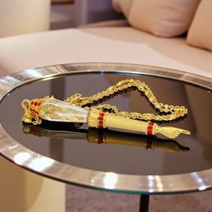 May include: A gold-colored necklace with a pendant featuring a pen-like design. The pendant has a white and gold body with red accents and a gold chain. The necklace is displayed on a reflective black surface.