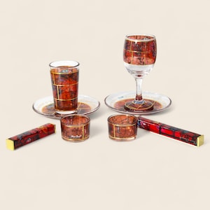 Op de afbeelding: Een verzameling glaswerk en accessoires met een abstract rood en goud patroon. Inclusief een wijnglas, borrelglas, schotels en kleine kaarshouders met gouden randen.