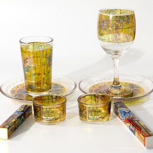 Op de afbeelding: Een verzameling glaswerk en decoratieve items met een glas-in-lood ontwerp. Inclusief een wijnglas, een drinkglas, twee kleine kaarshouders, twee borden en twee decoratieve dozen. Het glaswerk heeft een gouden rand.