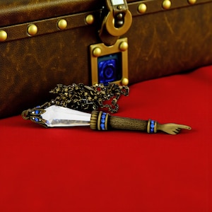 May include: A steampunk pendant necklace featuring a clear crystal pendant and a bronze chain. The pendant has blue accents and a hand-shaped pointer. A brown leather chest is in the background.