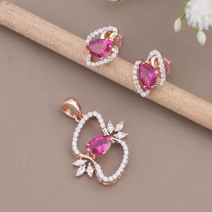 May include: A jewelry set consisting of a pendant and two stud earrings in rose gold. Each piece features a central kite-shaped fuchsia gemstone surrounded by a delicate frame of small white pavé stones, with additional marquise-cut accents.