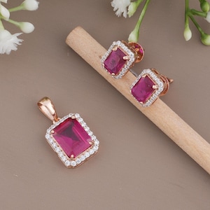 May include: A jewelry set featuring a pendant and matching stud earrings. Each piece has a rectangular, deep pink gemstone surrounded by a halo of small, clear stones. The metal has a rose gold finish.
