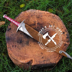 May include: A decorative sword with a red and silver handle and a brown sheath, resting on a wooden surface. The sword has intricate silver detailing and is set against a backdrop of green grass. The text "UMAR'S ART WORK" is visible.