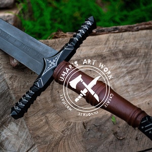 May include: A detailed image of a black and silver sword with a brown handle, resting on a wooden surface. The sword features intricate designs and inscriptions on the blade. The handle has a leather-like appearance. The image includes a logo that says "UMAR'S ART WORK".