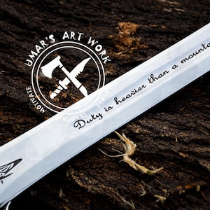 May include: A silver knife with a black handle and a white inscription that reads "Duty is heavier than a mountain". The knife is on a brown wooden surface. The inscription is in a cursive font. The knife has a black and white logo on the handle that reads "Jumar's Art Work".