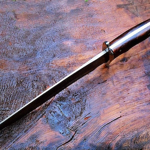 Puede incluir: Un cuchillo artesanal con un mango de madera oscura y una hoja larga y delgada. El mango presenta una franja de color claro en el centro. El cuchillo est&aacute; sobre una superficie de madera texturizada.