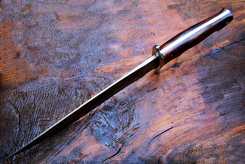 Puede incluir: Un cuchillo artesanal con un mango de madera oscura y una hoja larga y delgada. El mango presenta una franja de color claro en el centro. El cuchillo est&aacute; sobre una superficie de madera texturizada.