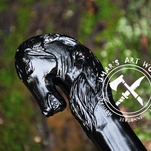 May include: A black wooden walking stick with a carved horse head handle. The horse head is detailed with a mane and a mouth with teeth. The stick is polished and has a smooth finish. The text "UMAR'S ART WORK CREATE INNOVATE MOTIVATE" is printed on the stick.