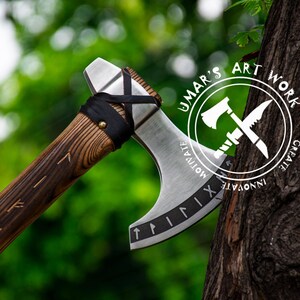 Op de afbeelding: Een handgesmede bijl met een houten steel en een metalen kop. De kop is versierd met Romeinse cijfers en een leren wikkeling. De bijl rust tegen een boomstam. De boomstam heeft een wit logo met de tekst "UMAR'S ART WORK CREATE INNOVATE MOTIVATE".