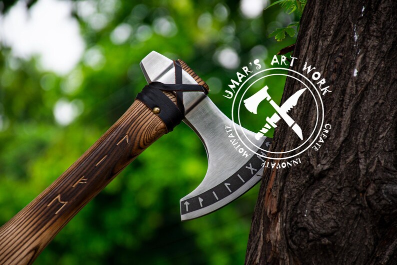 Op de afbeelding: Een handgesmede bijl met een houten steel en een metalen kop. De kop is versierd met Romeinse cijfers en een leren wikkeling. De bijl rust tegen een boomstam. De boomstam heeft een wit logo met de tekst "UMAR'S ART WORK CREATE INNOVATE MOTIVATE".