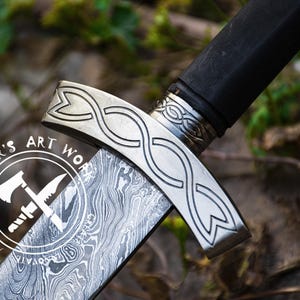 May include: Close-up of a silver sword with a black handle and a decorative knotwork design. The blade has a patterned finish and the hilt has the words "UMAR'S ART WORK" and "MOTIVATE" engraved on it.