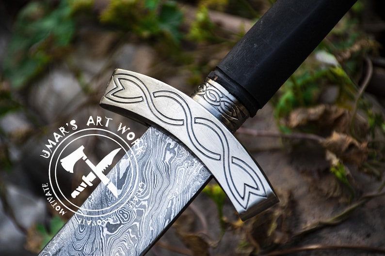 May include: Close-up of a silver sword with a black handle and a decorative knotwork design. The blade has a patterned finish and the hilt has the words "UMAR'S ART WORK" and "MOTIVATE" engraved on it.