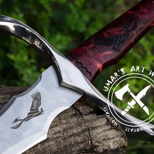 May include: A silver sword with a red and black wooden handle. The blade has an etched design of a bird in flight. The handle has a carved bird design. The sword is resting on a wooden surface. The sword has a silver guard and pommel. The sword is a decorative piece and is not intended for use as a weapon. The sword has the words "UMAR'S ART WORK CREATE MOTIVATE INNOVATE" etched on the handle.