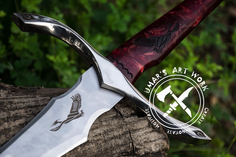 May include: A silver sword with a red and black wooden handle. The blade has an etched design of a bird in flight. The handle has a carved bird design. The sword is resting on a wooden surface. The sword has a silver guard and pommel. The sword is a decorative piece and is not intended for use as a weapon. The sword has the words "UMAR'S ART WORK CREATE MOTIVATE INNOVATE" etched on the handle.