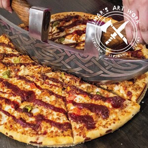 May include: A stainless steel pizza cutter with a curved blade featuring an intricate Celtic knotwork cutout design and two wooden handles. The blade is positioned over a pizza topped with red sauce, cheese, and peppers. The logo UMAR'S ART WORK is visible.