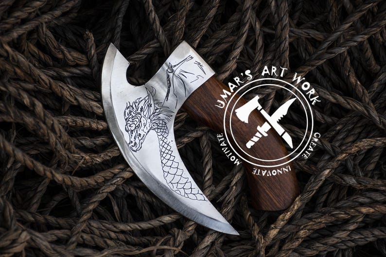 Op de afbeelding: Een zilveren bijl met een drakenontwerp op de kling. De bijl steel is gemaakt van hout en heeft een rond logo met de tekst "UMAR'S ART WORK CREATE INNOVATE MOTIVATE".