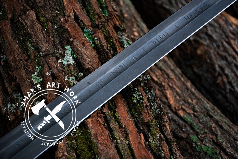 May include: A black sword with etched inscriptions, resting on a textured wood surface. The sword's blade is dark, and the handle is not visible. The image includes a white circular logo with the text "UMAR'S ART WORK".