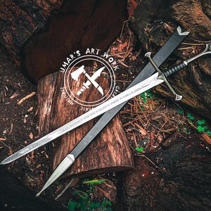 May include: A silver sword with a black handle and a black sheath rests on a wooden surface. The sword has intricate designs on the blade and handle. The image also features a logo with the text "UMAR'S ART WORK".