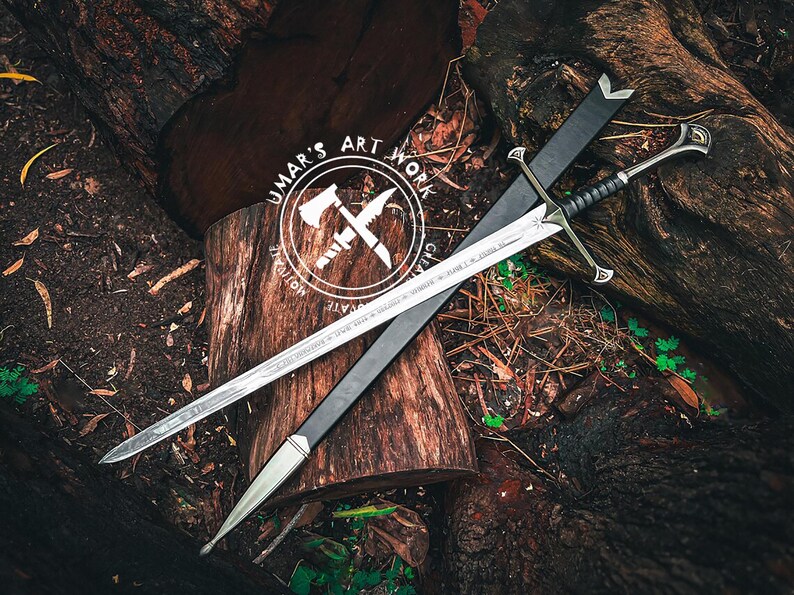 May include: A silver sword with a black handle and a black sheath rests on a wooden surface. The sword has intricate designs on the blade and handle. The image also features a logo with the text "UMAR'S ART WORK".