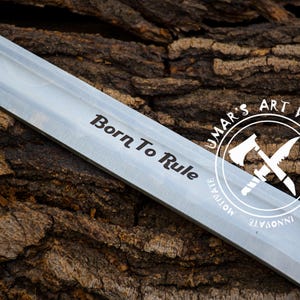 May include: Close-up of a silver blade with the words "Born To Rule" engraved on it. The blade is resting on a piece of tree bark. A circular logo with the text "UMAR'S ART WORK" is visible in the background.