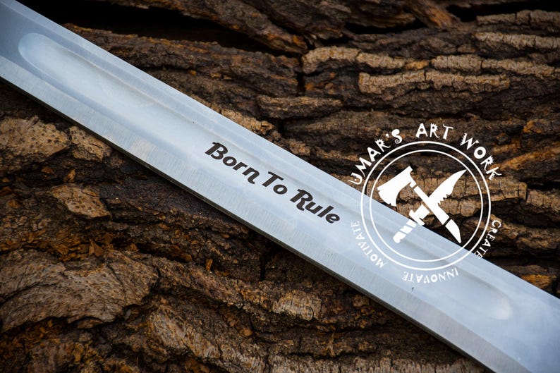 May include: Close-up of a silver blade with the words "Born To Rule" engraved on it. The blade is resting on a piece of tree bark. A circular logo with the text "UMAR'S ART WORK" is visible in the background.