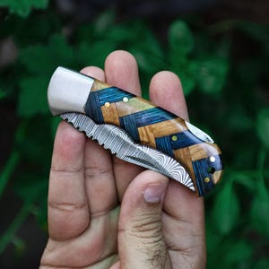 Puede incluir: Un cuchillo plegable de bolsillo con mango de madera azul, verde y marrón y una hoja de acero damasco.