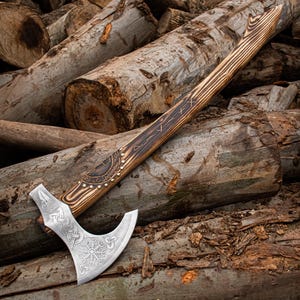 Puede incluir: Un hacha vikinga forjada a mano con un mango de madera y tallas intrincadas. La cabeza del hacha está pulida y presenta un diseño rúnico. El hacha está apoyada en una pila de troncos.