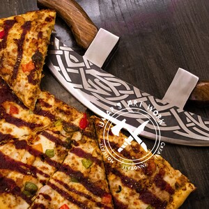 May include: A curved mezzaluna pizza cutter with a dark wood handle and a silver blade featuring an intricate etched Celtic knot pattern, positioned next to a sliced pizza with red sauce, cheese, and peppers. The blade displays the text UAAAR'S ART WORK.