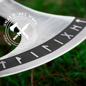 Op de afbeelding: Een zilveren bijl met een zwarte steel en een zwarte band rond de kling. De band heeft Romeinse cijfers erop. De bijl heeft een logo aan de zijkant dat "Umar's Art Work" zegt met een gekruist bijl- en mesontwerp. Het logo zegt ook "Motivate, Innovate, Create".
