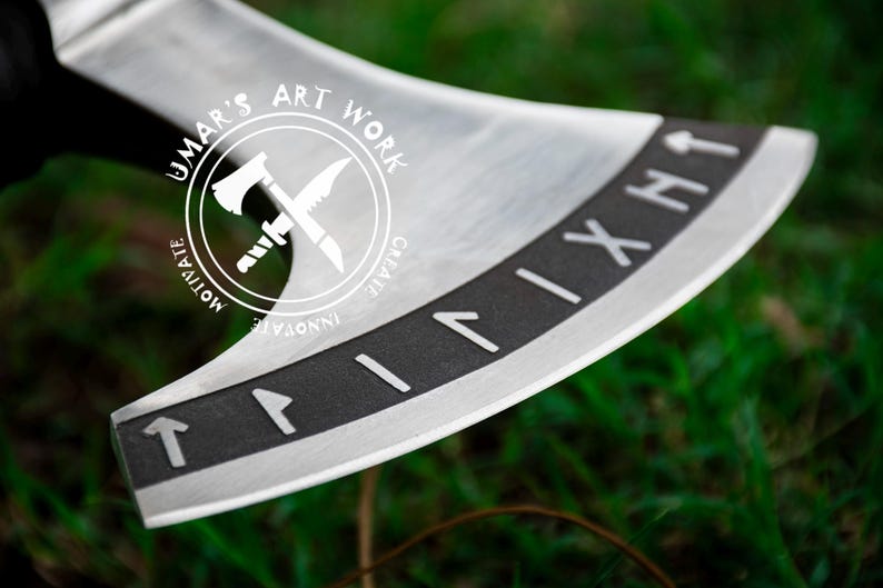 Op de afbeelding: Een zilveren bijl met een zwarte steel en een zwarte band rond de kling. De band heeft Romeinse cijfers erop. De bijl heeft een logo aan de zijkant dat "Umar's Art Work" zegt met een gekruist bijl- en mesontwerp. Het logo zegt ook "Motivate, Innovate, Create".