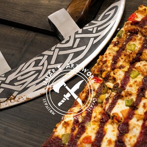 May include: A curved metal pizza cutter with a dark wooden handle and an intricate etched Celtic knot pattern on the blade. The blade features a circular logo with the text UMAR'S ARTWORK, MOTIVATE, INNOVATE, CREATE. A pizza with toppings sits nearby.