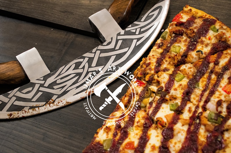 May include: A curved metal pizza cutter with a dark wooden handle and an intricate etched Celtic knot pattern on the blade. The blade features a circular logo with the text UMAR'S ARTWORK, MOTIVATE, INNOVATE, CREATE. A pizza with toppings sits nearby.