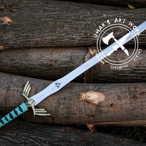 May include: A silver-colored sword with a teal and gold handle, resting on a pile of logs. The sword features a triangular emblem and gold accents. The handle is wrapped in a teal and black pattern. The image includes a circular logo with text.